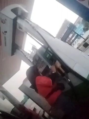 A person wearing a red jacket and dark pants is standing next to a white gas pump. The person appears to be interacting with the gas pump or its components, possibly filling a vehicle. The lighting suggests it is daytime, and the environment looks like a gas station. The video is shaky and at an angle, making it difficult to discern fine details.