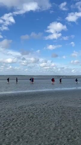 A group of people are walking across a shallow body of water on a sandy shore. The sky is blue with scattered white clouds. The people are spread out, some walking alone and others in small groups. The water is calm and reflects the sky. The sand is wet and appears dark, indicating it has recently been submerged by water. The overall scene suggests a coastal environment during daylight hours.