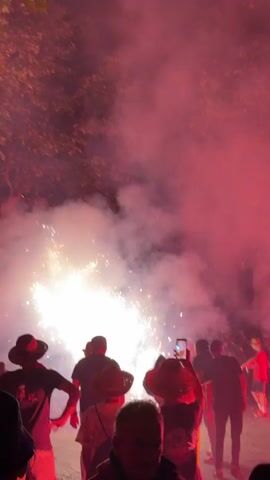 A crowd of people watch fireworks. The fireworks are bright white and yellow sparks exploding from a central point, creating a large amount of smoke that is illuminated by red and pink light. Many people are wearing hats and some are holding up their phones to record the event. The scene appears to be outdoors at night.