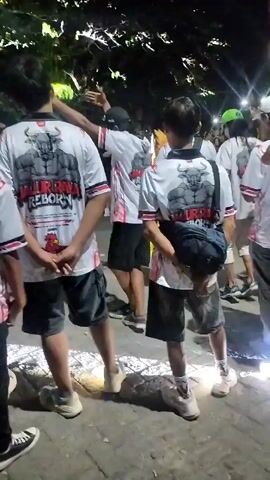 The video shows a group of people from the back at night. They are wearing matching white t-shirts with a graphic of a muscular bull and the words "MAJU REBORN" on the back. Some of the individuals are raising their hands in the air. In the background, there are lights and trees, suggesting an outdoor setting. The ground appears to be paved. The overall atmosphere seems to be celebratory or part of an event.