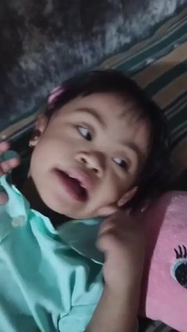 The video shows a young child lying on their back on what appears to be a striped bed or cushion. They are wearing a light blue button-down shirt. A pink plush toy is beside the child. An adult's hand is briefly seen touching the child's cheek. The child looks directly at the camera and opens their mouth slightly. The lighting is dim.
