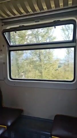 The video shows the interior of a train carriage, with seats visible in the foreground. A window looks out onto a blurry landscape of trees and sky, indicating the train is in motion. The window is partially open, allowing in light and air. The interior of the train appears to be beige and brown, with some yellow accents on the seats. The overall lighting suggests it is daytime.