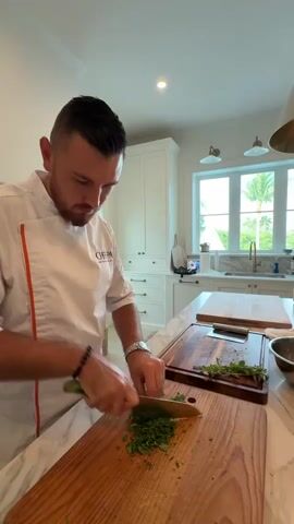 A chef in a white uniform with orange trim is chopping green herbs on a wooden cutting board. He is focused on the task, holding a knife in his right hand and guiding the herbs with his left. The kitchen is bright and modern, with white cabinets, a marble countertop, and a large window in the background that reveals palm trees and a blue sky. Other kitchen tools and ingredients are visible on the counter, including another cutting board with herbs and a bowl. The overall impression is one of a professional chef preparing ingredients in a clean and well-lit kitchen.
