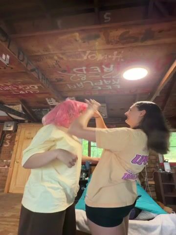 A young person with short pink hair, wearing a yellow t-shirt and brown pants, stands on the left side of the frame, their hands raised to their head. Facing them is another young person, wearing a peach t-shirt and black shorts, with long dark hair. They are on the right side of the frame, also with their hands near the pink-haired person's head. They appear to be interacting, possibly playing or styling the pink hair. The background suggests an indoor setting, possibly a cabin, with a wooden ceiling covered in graffiti. A window is visible on the right, showing greenery outside. A bed with blue and green bedding is also in the background.