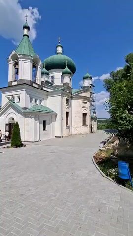The video begins with a view of a white church with green domes under a bright blue sky with some clouds. The church has a bell tower. People are visible standing near the entrance of the church and on the grounds. The camera pans to the left, showing more of the church's facade and the surrounding paved area. The camera then moves further left, revealing another building that appears to be part of the church complex, made of stone. More people are seen walking on the grounds and near the entrance of the church. The camera continues to pan, showing a shaded area with trees and a curved pathway. The scene is set in an outdoor environment with a clear sky and green foliage, suggesting it is daytime and likely pleasant weather. There are some construction elements visible on the side of the church, possibly scaffolding. A blue trash receptacle is also visible.