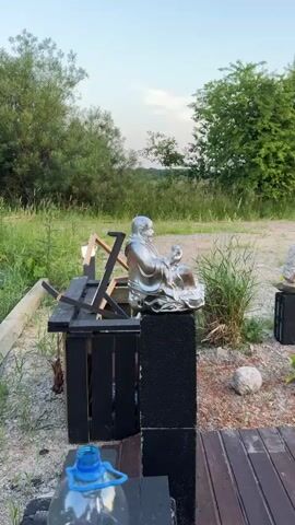 The video shows an outdoor scene with a clear blue sky. In the foreground, there is a silver statue of a seated figure, possibly a Buddha, placed on a black pedestal. To the left of the statue, there is a black wooden structure with metal supports, resembling a bench or a stand. Partially visible in the foreground left is an orange plastic bag. In the background, there is lush green grass and trees. The overall setting appears to be a yard or an open outdoor space, possibly at dusk or dawn given the lighting.