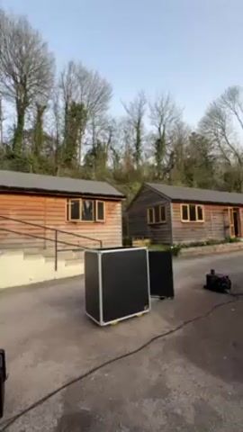 A camera films two small, wooden buildings on a lot covered with stones. Between them, a black, boxlike machine spews two streams of fire upward into the air, and then stops. There are bare trees behind the buildings and a light blue sky above.