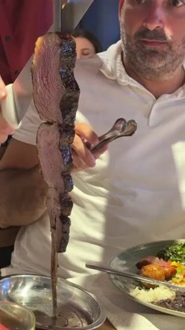 A man in a white polo shirt is carving a large piece of meat on a skewer. The meat appears to be a roast, with a thick layer of fat on top. He is holding a knife in his right hand and the skewer in his left. The skewer is placed on a metal stand, which is on a table. There is a plate of food in the foreground, with rice, beans, and green vegetables. The background is blurred, but it appears to be a restaurant setting.