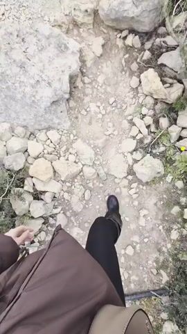 A person wearing black leggings and black boots is walking along a rocky path.  A second person wearing dark pants and white sneakers passes, walking over rocks. The path is uneven and covered in loose gravel and small rocks, bordered by larger rocks and sparse vegetation.  The sky is overcast. The camera angle is a first-person perspective, focusing on the path and the person walking.