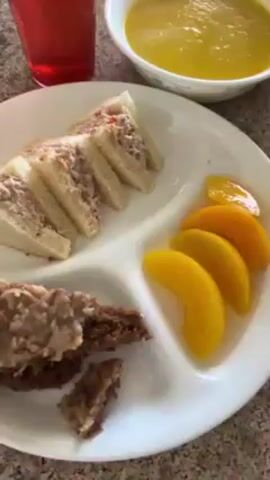 The video shows a close-up of a table set with several plates of food, glasses of drinks, and bowls of soup. The plates contain sandwiches cut into triangles, slices of peaches, and pieces of cake. The drinks appear to be water and a red-colored beverage with ice. The soup in the bowls is yellow. The camera pans slightly to show multiple place settings with similar food arrangements. The table is a light brown color with a speckled pattern. The overall setting seems to be a casual meal, possibly lunch or a light supper.