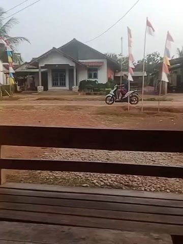 A short video shows a residential street with several houses and some vegetation. In the foreground, a wooden bench is visible. In the middle ground, a person on a white motorcycle is riding past. In the background, there are houses, some with flags displayed, and trees. The ground is covered with dirt and some grass. The sky is overcast.