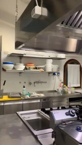 The video shows a panning shot of a professional kitchen. The camera moves from left to right, revealing various kitchen equipment. On the left, a stainless steel sink and a commercial stove with a fryer are visible. Above the stove, a large stainless steel exhaust hood is installed. The kitchen walls are tiled, and shelves are mounted on them, holding various kitchenware and cleaning supplies. To the right, a stainless steel countertop houses a commercial oven and other appliances. A window with a white curtain is visible in the background. Further to the right, a commercial meat slicer and a microwave are on a countertop. The floor is tiled. The overall impression is of a clean and well-equipped commercial kitchen.