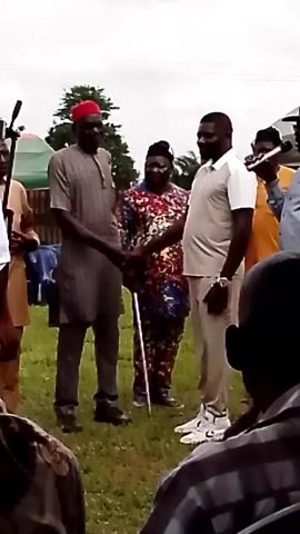The video shows a group of people outdoors, seemingly at an event. Two men in the foreground are shaking hands. The man on the left wears a grey traditional outfit and a red cap, while the man on the right wears a light-colored polo shirt and pants. A person in a colorful patterned outfit stands between them, holding a cane. Other individuals are visible in the background, some holding microphones. The environment appears to be a grassy area with trees and some structures in the distance.