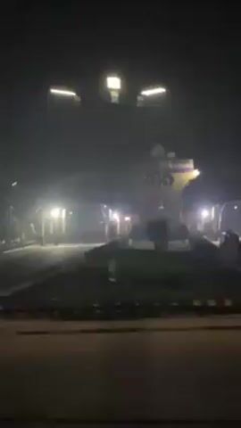 The video, taken from a moving vehicle at night, shows an illuminated archway with text on it and what appears to be a military tank in front of it. The archway is surrounded by streetlights and appears to be on a wide, possibly grassy median strip of a road. The video then pans to the right, showing trees and a dark, undefined background behind the archway. Visibility is low due to the darkness. The camera movement is smooth, indicating it is likely being recorded from a car or bus window. No people are clearly visible in the video.