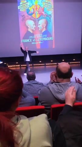 The video shows a man on stage taking a bow and clapping in front of an audience in a theater. The man is bald and wearing a gray suit jacket, a white shirt, and gray pants. The audience is seated in red chairs and applauding. Behind the man on stage is a large screen displaying a poster that says "JULIEN STRELZYK FRANCE vs LUXEMBOURG."  The poster has images of two bald men facing each other, with a rooster and a lion above their heads. There are stage lights along the front of the stage. The camera is positioned from the audience's perspective.