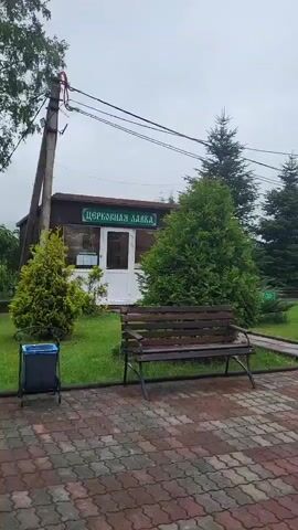 A person walks down stairs outside a brick building. The building has a green roof and a gold cross on top. There are trees and grass surrounding the building. The ground is paved with red and gray stones. A wooden bench is visible in the foreground. There is a sign above a door that reads "ЦЕРКОВНАЯ ЛАВКА". The sky is overcast.