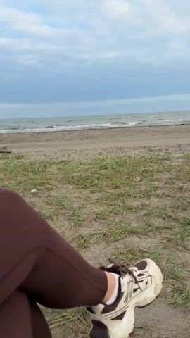 The video shows a close-up view of a person's legs and feet sitting on a sandy beach. The person is wearing brown leggings and athletic shoes with white socks. The background features the ocean, a sandy shore, and green grass. The sky is cloudy. The camera is positioned at a low angle, focusing on the person's lower body and the beach landscape.