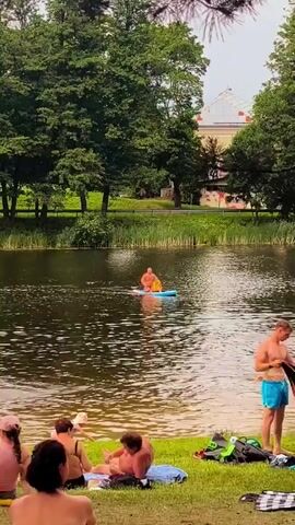 The video shows a lake on a sunny day. Several people are sunbathing on the grassy shore, and some are in the water. One person is on a paddleboard in the middle of the lake. A man is standing on the shore holding some items. There are trees and a building in the background.