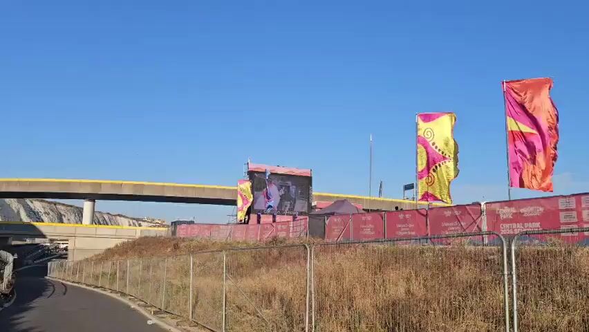 The video shows an outdoor event. A large screen displays a video of a person in blue and orange clothing holding a flag. Two colorful flags, one yellow with pink designs and another orange and pink, wave in the wind. A yellow overpass crosses the background, and a clear blue sky is visible above. Red fencing with white text encloses the event area, and a purple tent is set up behind the screen. Dry grass and a wire fence are in the foreground.