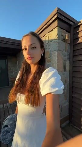 The video shows a young woman standing on a wooden deck in front of a building with stone and wooden accents. She is wearing a white puff-sleeve dress and holding a patterned scarf in her left hand. The sun is casting a warm glow, indicating it is either sunrise or sunset. The sky is clear and blue. The woman has long, brown, wavy hair and is looking directly at the camera with a slight smile. Her eyes are closed in some frames, and open in others. She is wearing a necklace and earrings. In the background, there are wooden shutters on the building, and some outdoor furniture, suggesting a vacation or resort setting. The overall mood is relaxed and peaceful. No text is visible in the video.