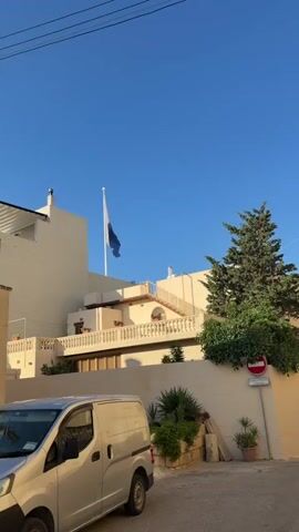 The video shows a light blue sky with a white and blue flag blowing in the wind on a flagpole. The flag is on a building with beige walls and a white railing. There are stairs leading up to a balcony with potted plants. A green tree is visible on the right side of the frame. The bottom of the frame shows the top of a white vehicle and a wall.