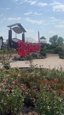 The video opens with a close-up shot of a large, bright pink sign with Cyrillic lettering, likely a place name. In the background, there is an archway with a smaller sign above it, and beyond that, a building and a wooden structure resembling a gazebo or observation deck. Lush green plants and colorful flowers are in the foreground, obscuring the base of the sign. The camera then pans to the right, revealing more of the landscape. A paved pathway leads towards figures standing in the distance. A horse-drawn cart is visible on the pathway, with what appear to be figures dressed in historical costumes standing nearby. Further down the path, two people are seen talking. The landscape transitions to a more open, arid area with a church-like structure on a hill in the background. The video concludes with a shot of a horse harnessed to a carriage, with several more carriages lined up behind it. The environment appears to be an outdoor recreational area or historical site, with elements suggesting a rural or agricultural theme. The sky is clear with scattered clouds, and the lighting suggests it is daytime.