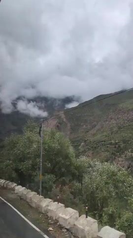 The video is taken from a moving vehicle, looking out a window at a mountainous landscape under a cloudy sky. The road is visible in the foreground, with a concrete barrier along its edge. A utility pole with wires is positioned on the right side of the frame. The mountains in the background are covered in vegetation and appear rugged, with some exposed rock faces. Clouds are present, obscuring parts of the upper mountain peaks. The overall atmosphere is somewhat overcast and possibly damp, suggesting a cooler climate or recent rain.