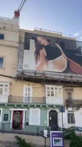 The video shows a view from the top of an open-top tourist bus in what appears to be a coastal city. The bus is moving along a road next to a body of water. Buildings of various colors and architectural styles line the street. Some buildings are older with balconies, while others are modern apartment blocks. Several shops and businesses are visible at street level. A red and white biplane flies overhead. People are seated on the bus. The sky is clear and blue.