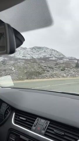 The video captures a first-person perspective from the interior of a car driving through a wintry, mountainous landscape in Provalija, Montenegro. The dashboard of the car is prominent in the foreground, with the rearview mirror visible at the top of the frame.

Through the windshield, a two-lane road stretches ahead, bordered by a metal guardrail. Beyond the guardrail, the terrain is rugged and rocky, with the ground and numerous boulders lightly dusted with snow. In the background, large mountains covered in a more substantial layer of snow rise under a uniformly overcast, grey sky. Sparse, low-lying trees, some coniferous, are scattered across the rocky landscape, also bearing a light covering of snow. The overall impression is one of a stark and cold, yet scenic, winter environment.

Inside the car, the black dashboard is visible, featuring air vents and a central console with a red hazard light button. Text below the hazard button reads "PASSENGER AIRBAG". A partial view of the instrument cluster shows digits, with "176" visible, likely indicating speed in km/h or another numerical reading. The car is in motion, steadily traversing the mountain road. The time of day appears to be daytime, though the heavy cloud cover gives the scene a muted light.