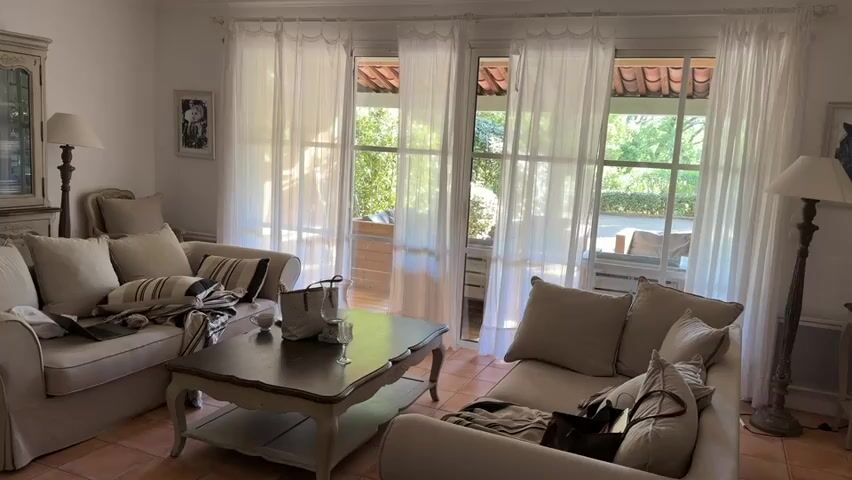 The camera slowly pans from left to right across the interior of a room. The room is furnished with two sofas and a coffee table with a glass and tote bag on it. The sofas are arranged facing each other. The walls are white and have a painting. Two lamps are also visible in the room. Large windows with sheer white curtains open onto an outdoor patio area with outdoor furniture.  The floor is tiled. The room appears to be well-lit and casually decorated.
