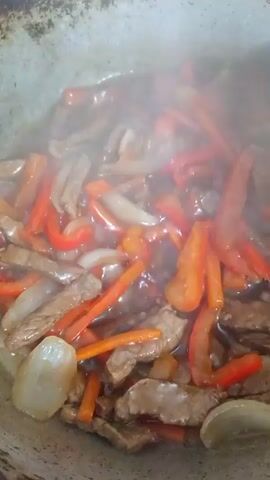 A close-up shot of a stir-fry dish being cooked in a wok. The dish contains strips of meat, sliced carrots, onions, and red bell peppers. Steam is rising from the wok, indicating that the food is hot. A metal utensil, possibly a spatula or tongs, is used to stir the ingredients. The camera is positioned above the wok, looking down into the cooking food. The lighting is natural and appears to be from an overhead source. The background is blurred, focusing attention on the food.