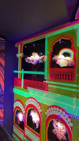 The video pans across a vibrantly lit, multi-story structure. The structure is made of translucent, brightly colored materials, predominantly neon green and hot pink. It features architectural elements reminiscent of Indian palaces, with arched windows and decorative railings. Inside some of the arched openings, illuminated objects are visible, including what appears to be a winged heart, a rainbow-like arc over a globe, and various intricate, light-emitting sculptures. The entire structure is adorned with strings of small, colorful lights that twinkle and pulse. The lighting creates a surreal and festive atmosphere, suggesting an art installation or a decorated building. The camera angle is from a slightly low perspective, looking up at the structure, and the lighting is intense, causing some chromatic aberration and blurring.