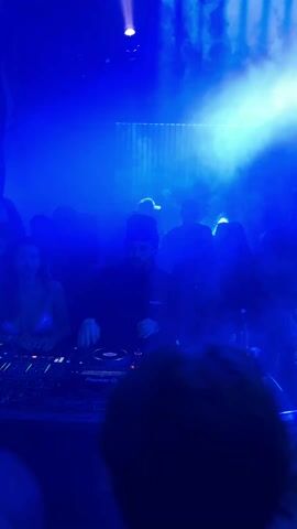 The scene appears to be inside a nightclub or music venue. The predominant lighting is blue, with some purplish hues and spotlight effects. A DJ is standing behind a mixing console with equipment, facing the crowd. There are a number of people in the background dancing and moving around in the dimly lit space, some are visible only as silhouettes. The DJ is wearing a dark-colored cap and clothing. A woman wearing a light-colored top is visible to the left of the DJ. In the foreground, the back of a person's head with short hair and glasses is visible. Cables and wires from the DJ equipment are scattered on the table. The backdrop includes vertical textured panels illuminated by the blue light. There appears to be some kind of smoke or haze in the air.