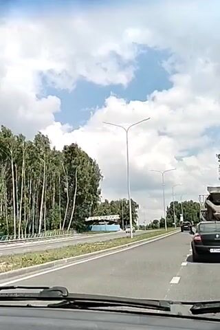 A dashcam view shows a car driving on a road. A line of birch trees is on the left side of the road. A blue circular structure with white lettering is visible beyond the trees. Several cars are on the road ahead, and a truck is on the right side of the road. The sky is blue with white clouds.