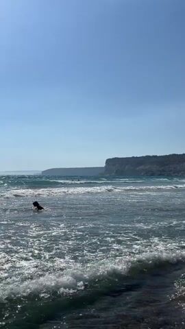 A first-person perspective video shows a person walking into the ocean on a sunny day. The water is clear and blue with small waves and white foam. In the distance, there are cliffs and a beach with a few people and umbrellas. The person's feet and legs are visible as they walk deeper into the water, which reaches their ankles and then their shins. The video ends with a close-up of the foamy water around the person's legs.