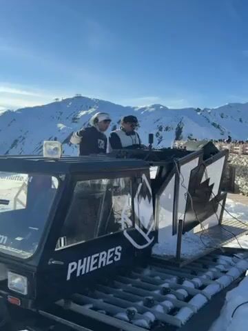 The video shows a party scene on a snowy mountain. Two DJs are on a snowcat labeled "PHIERES," with mountains in the background. The snowcat is rigged with speakers and a backdrop with the image of a maple leaf. People are dancing and socializing in the snow. The weather appears sunny. A building is visible in the background.