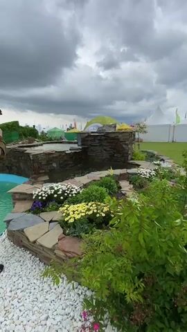 The video shows a stone pond with small waterfalls on a cloudy day, with event tents in the background. The pond is surrounded by landscaping including rocks, white pebbles, and flowering plants in white, yellow, and purple. The water in the pond is clear, and the camera appears to be stationary. A person's arm is briefly visible on the left side of the frame.