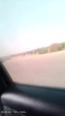 The video shows a car driving on a road. The landscape is dry, with sandy soil and scattered trees. A white Toyota pickup truck with writing on the side is parked on the roadside, with a man in a blue shirt visible in the passenger seat and a man wearing tan pants walking beside the truck. The sky is a hazy blue.