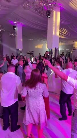 The video shows a crowded indoor event, likely a wedding reception, with a DJ booth visible in the background. People are dancing in a circle formation, with some individuals wearing formal attire, including a woman in a white wedding dress. The room is decorated with draped fabric ceilings and large spherical chandeliers. Colored lights, including purple and green, are projected onto the walls and dance floor, creating a vibrant atmosphere. Several people are holding hands and dancing in a line. The camera is handheld and appears to be moving with the crowd.