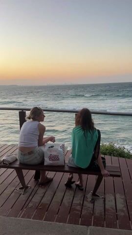 Two women are sitting on a wooden bench facing the ocean at sunset. The woman on the left is wearing a white tank top and light-colored shorts; the other woman has a green towel draped over her shoulders and is wearing shorts. A white shopping bag with the words "Salty Chook" printed in red is between them. The ocean is turquoise with visible waves, and the horizon is a gradient of yellow, orange, and pale blue. In the distance, a faint skyline is visible. The bench is on a wooden deck with a railing, and vegetation can be seen on the right side of the frame.