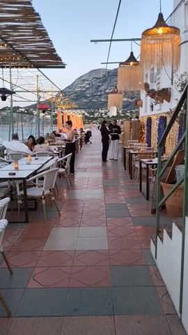The video shows a rooftop restaurant with a view of a mountain and the sky. Tables and chairs are arranged along a tiled walkway. Some tables have orange parasols. Straw lamps hang above. People are seated at some of the tables and others are walking along the walkway, including waitstaff.