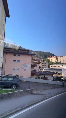 The video pans across a scenic view of a town nestled in a valley with mountains in the background under a clear blue sky. The town features buildings with terracotta roofs and pastel-colored facades, characteristic of European architecture. A prominent building in the foreground displays a sign that reads "Hotel Cristallo." Other structures, including what appears to be a modern building with large windows and solar panels, are visible. There are cars parked in a lot, and a road with a white line marking runs in the foreground. A street lamp and a bollard are also present. The general impression is that of a peaceful, sunny day in a picturesque mountain resort town, possibly during summer given the greenery and clear skies.