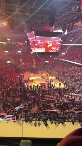 The video shows a high-angle view of a basketball court during a pre-game event. A large crowd fills the stands, and the court is illuminated with bright lights and spotlights.  The center court displays a graphic logo.  Several individuals in suits and team attire are on the court. A performer in a cowboy-style outfit dances and interacts with others, accompanied by music. There are visual effects of fire and pyrotechnics near the ceiling of the arena. A large video screen hangs above the court displaying images related to the event.