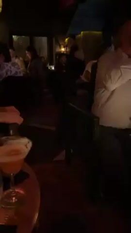 The video shows a crowded, dimly lit bar or restaurant. The ceiling is dark red. Several people are seated at tables, illuminated by flickering candlelight, while others stand and move around. A waiter carries a large tub over his head, navigating through the crowd. There is a staircase leading to another area. Decorative lamps hang from the ceiling. People are drinking and conversing, and some are holding up their phones, possibly recording the scene.