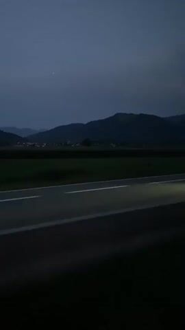 A video shows a road passing through a landscape of fields and hills under a dark, twilight sky. The road has white lines marking its lanes. On the right side of the road, a tall, skeletal power line tower is visible against the dusky sky. The scene suggests movement, likely from a vehicle traveling along the road. The overall atmosphere is dark and moody, typical of early morning or late evening.