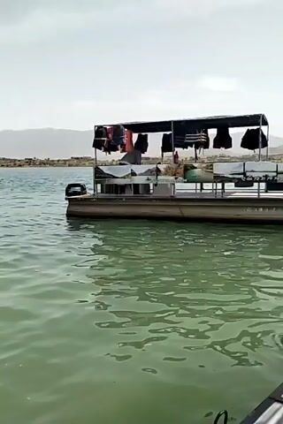 The video shows a stationary pontoon boat on a lake. The boat has a canopy structure on top with life vests hanging from it.  There are people visible on the boat. The water is calm and a greenish color. In the distance, there is a landscape of low-lying hills and a cloudy sky. The video is taken from a lower angle, likely from another boat on the water.