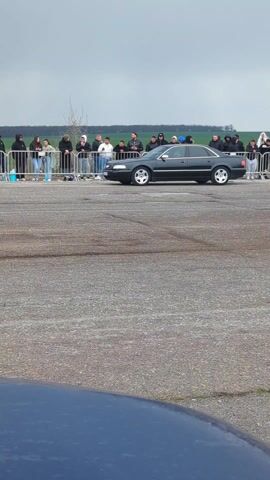 The video shows a dark gray sedan, possibly an Audi, parked on an asphalt surface. A crowd of people, mostly wearing dark hooded jackets, stands behind a metal barrier watching. The background includes a field with green vegetation and a cloudy sky. A portion of a blue vehicle is visible in the foreground.