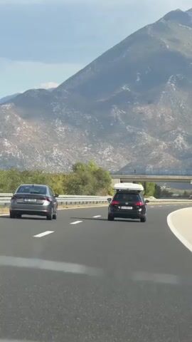 The video shows a view from inside a car driving on a highway. Two cars are visible ahead on the same lane, with a dark sedan in front and a black SUV with a roof rack behind it. The highway has white lane markings and a guardrail on the right side. In the background, there are mountains with sparse vegetation and a bridge crossing over the highway. The sky is partly cloudy with a bright sun. The camera is positioned to capture the road and the vehicles ahead.