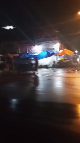 The video shows a nighttime street scene, possibly after rain. The ground is wet and reflective, with lights from streetlamps and buildings creating streaks and glows. Several vehicles are visible, including a red car on the left and a red bus-like vehicle in the center. There are temporary structures, possibly stalls or vendor tents, set up along the side of the road, some with blue and white tarps. The buildings in the background appear to be multi-story structures with signs and illuminated windows. A few indistinct figures can be seen, suggesting pedestrian activity. The overall impression is a lively, somewhat blurry urban environment at night, with elements of commerce and transportation.
