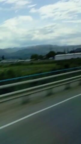 The video captures a view from a moving vehicle, likely a car or bus, traveling along a road. In the foreground, a metal guardrail separates the vehicle from a strip of green grass and foliage. Beyond this, several industrial-style buildings, possibly warehouses, are visible, some appearing white or light-colored. A blue truck and a yellow excavator can be seen near one of these buildings, suggesting some form of construction or industrial activity. Power lines and electricity pylons are present throughout the mid-ground. In the distance, a range of mountains stretches across the horizon under a sky filled with white and grey clouds. The scene depicts a daytime view in a rural or semi-urban area, possibly near an industrial zone, in Sayukovo, Russia. No visible text is legible.